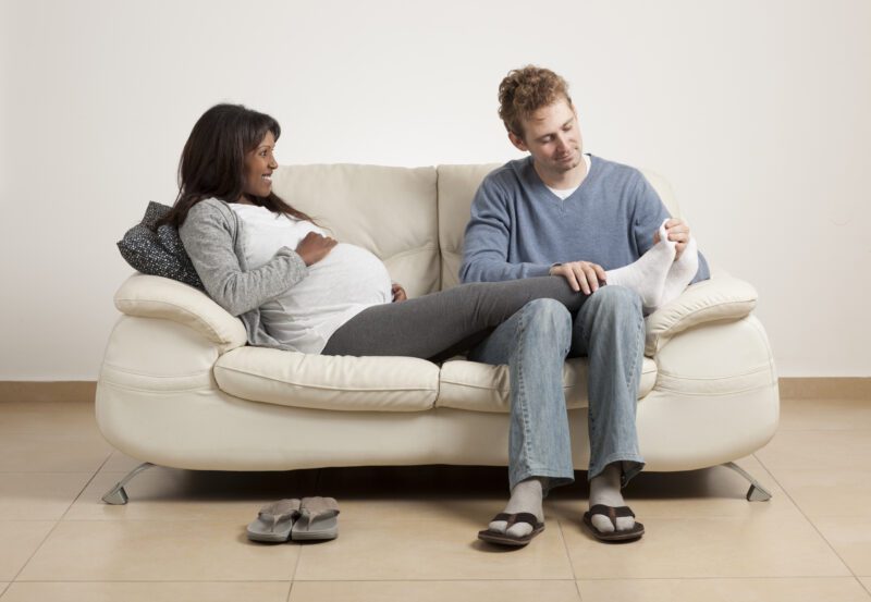 A young couple sitting on a couch. The woman is pregnant and the man is helping with foot pain. 