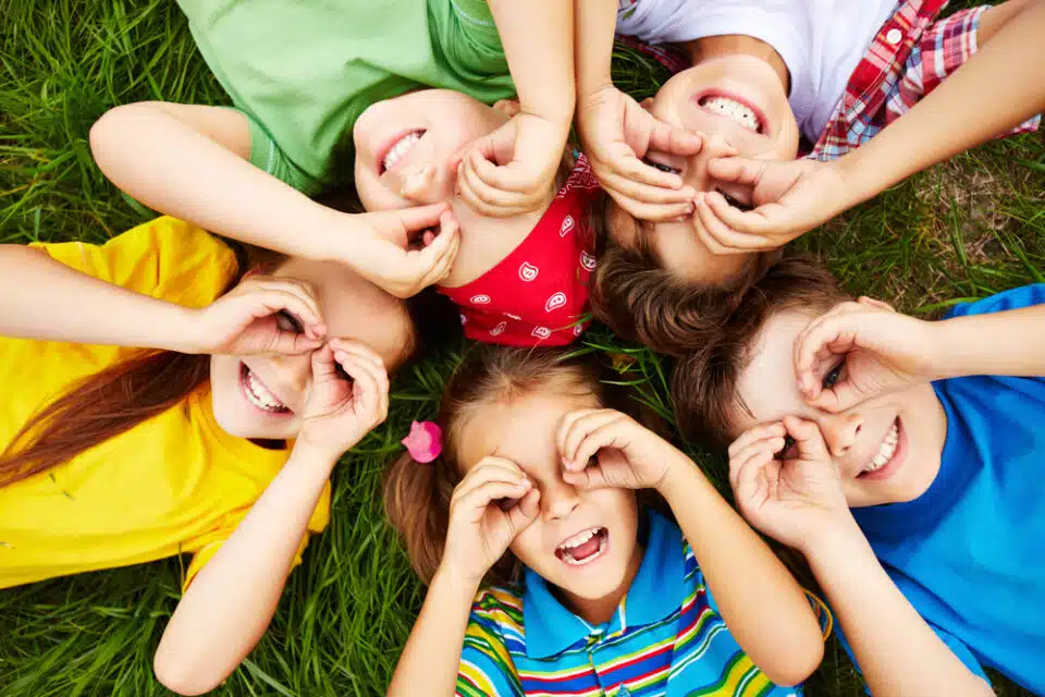 5 kids lying on the grass making a binoculars shape with their hands and smiling