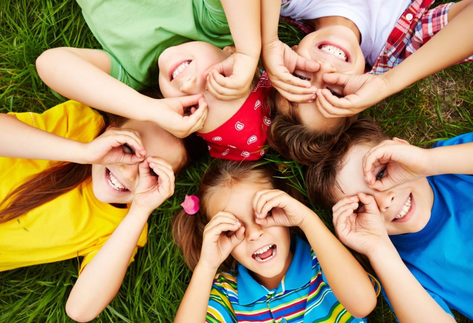 5 kids lying on the grass making a binoculars shape with their hands and smiling