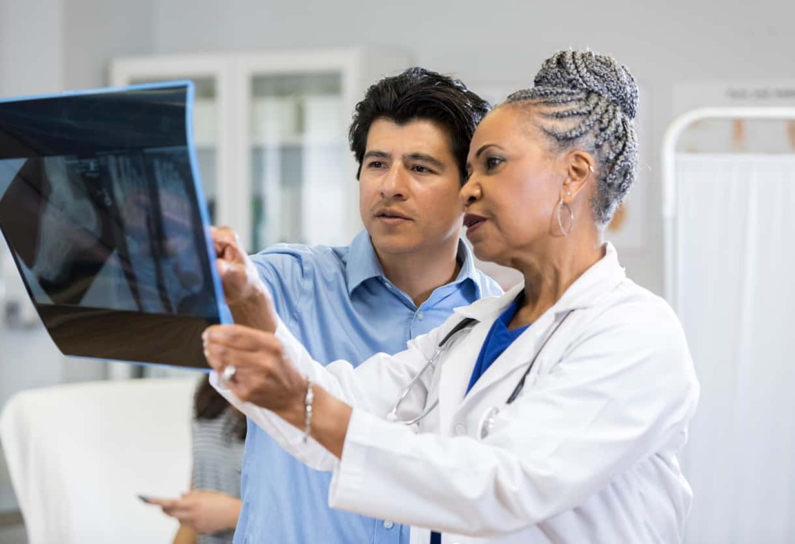 A concerned mid adult father points to a set of x-rays as a pediatrician shows him the images and shares the discovery.  They are standing in an exam room.  His teen daughter sits on a bed in the background.