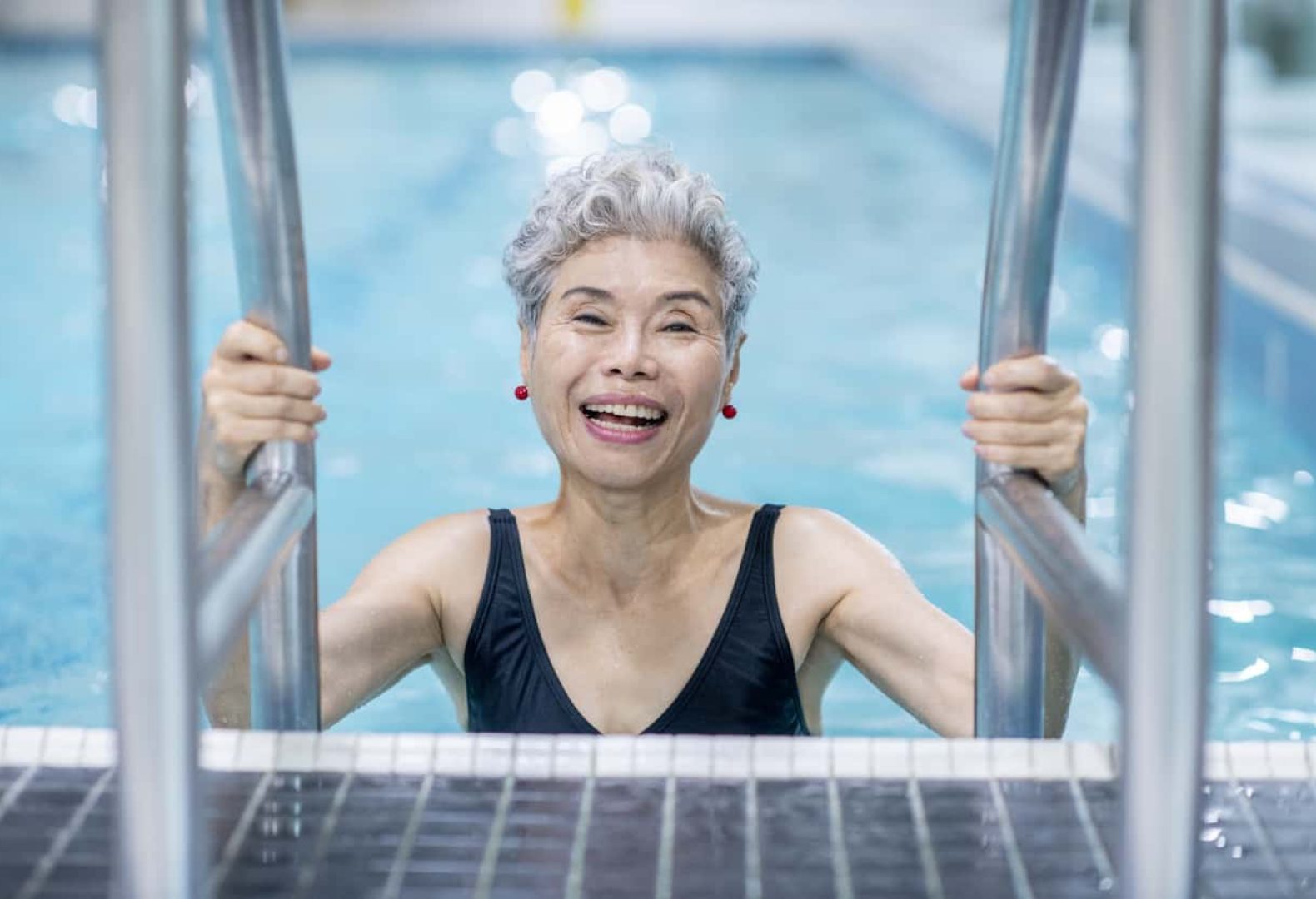 A senior Asian woman is at an indoor swimming pool. She is smiling at the camera while holding the ladder.
