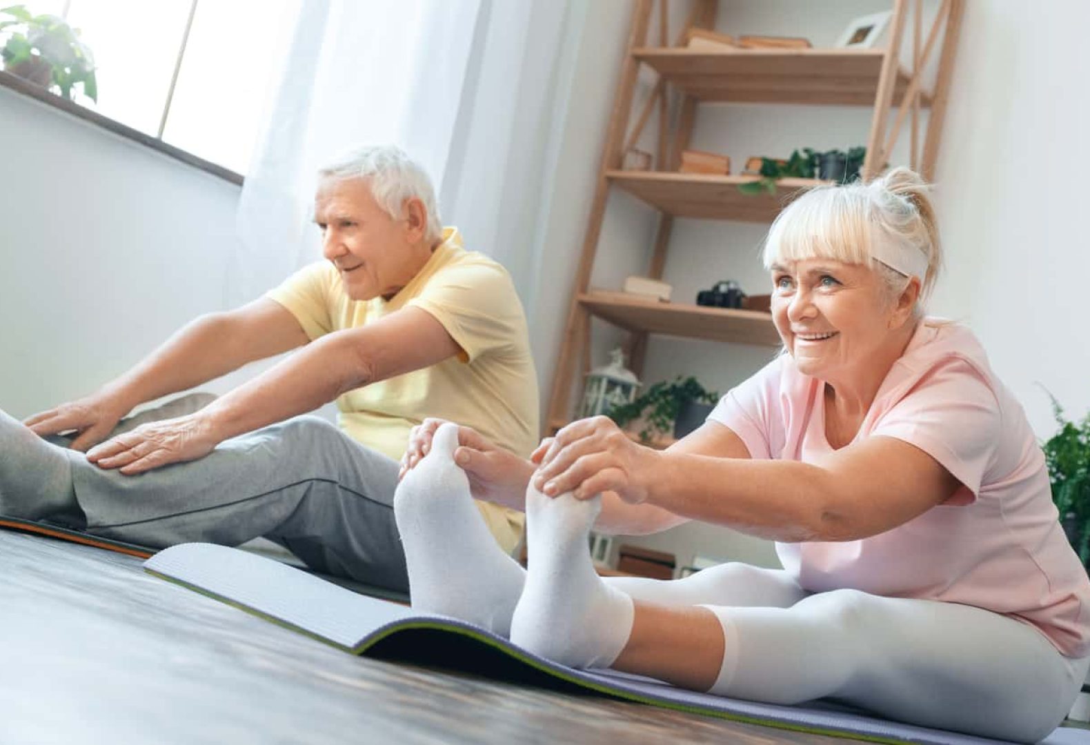 Senior man and woman doing yoga together indoors holding feet legs stretching smiling