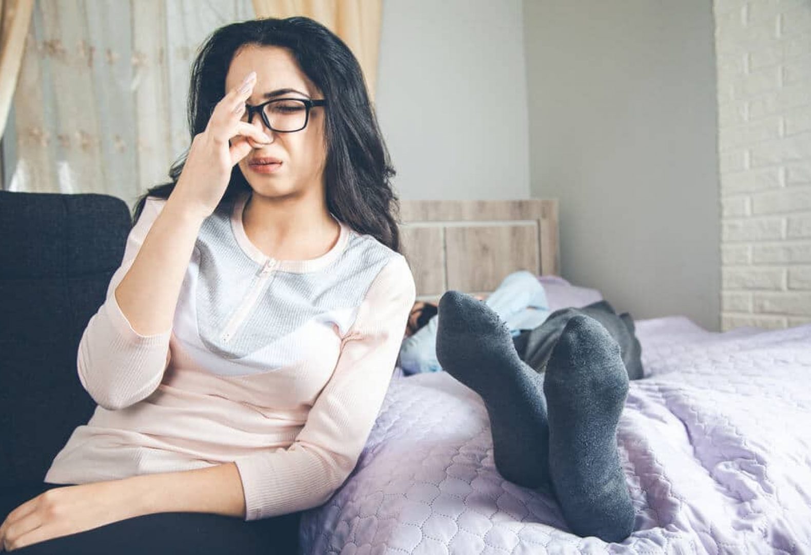 woman with glasses and dark hair pinching her nose due to stinky feet on bed next to her