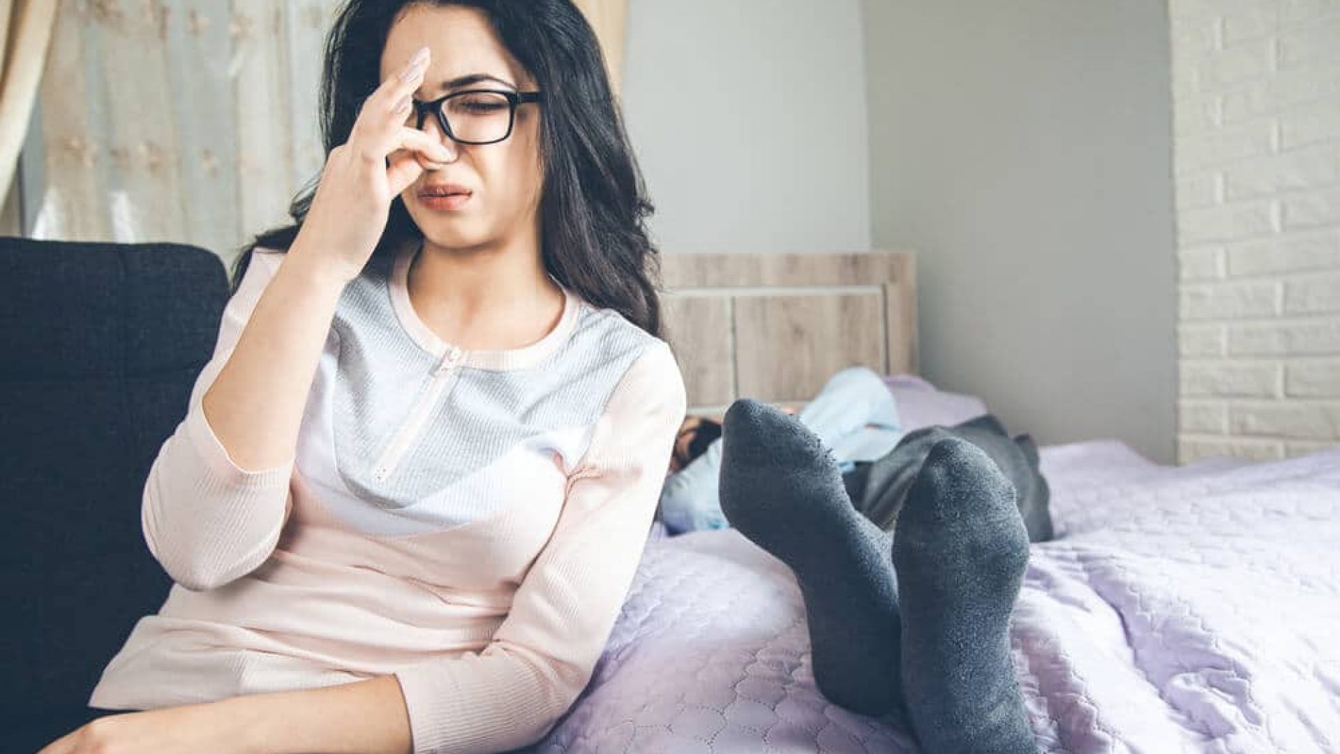 woman with glasses and dark hair pinching her nose due to stinky feet on bed next to her