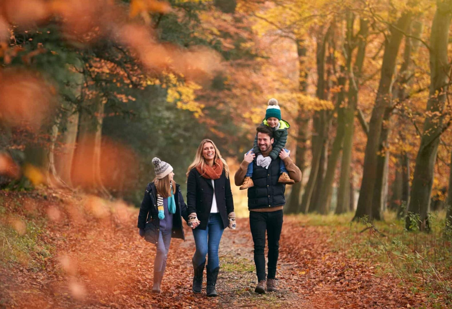 Family Walking Along Autumn Woodland Path With Father Carrying Son On Shoulders