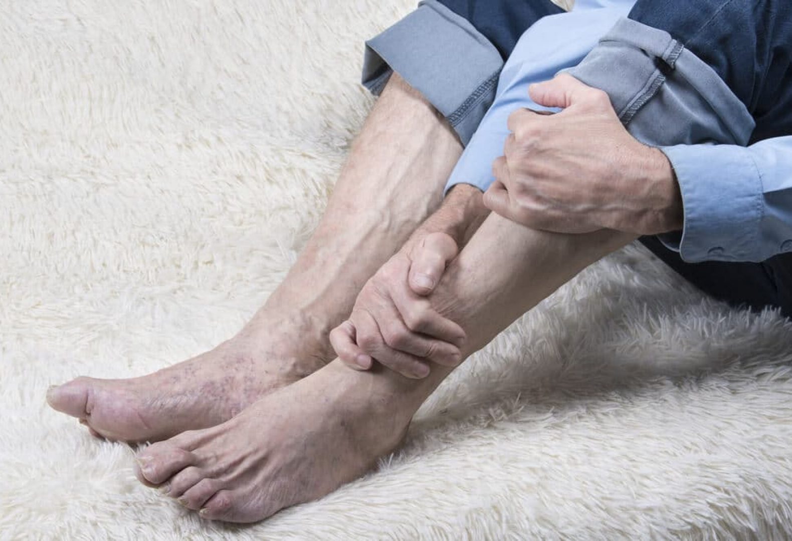 Man clutching his calf and lower leg with his pants rolled up on white carpet