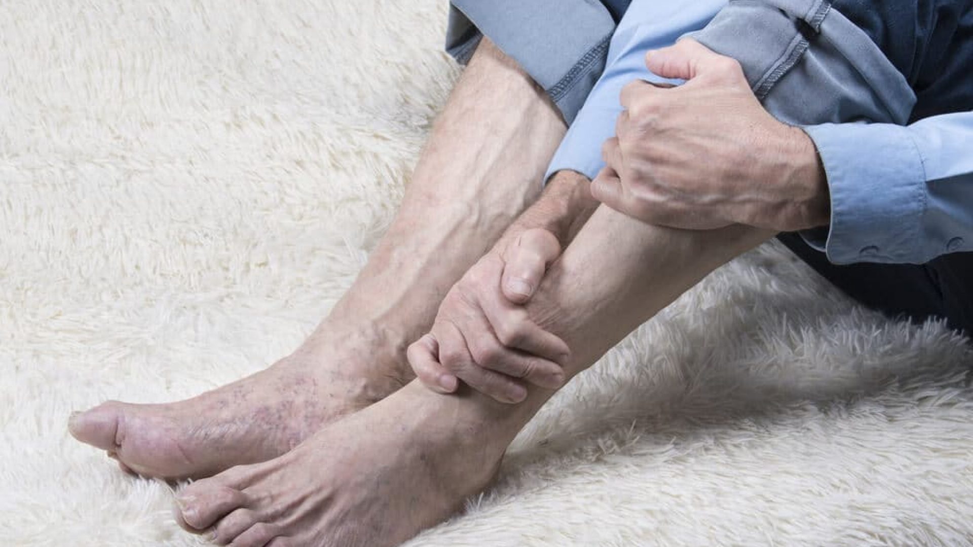 Man clutching his calf and lower leg with his pants rolled up on white carpet