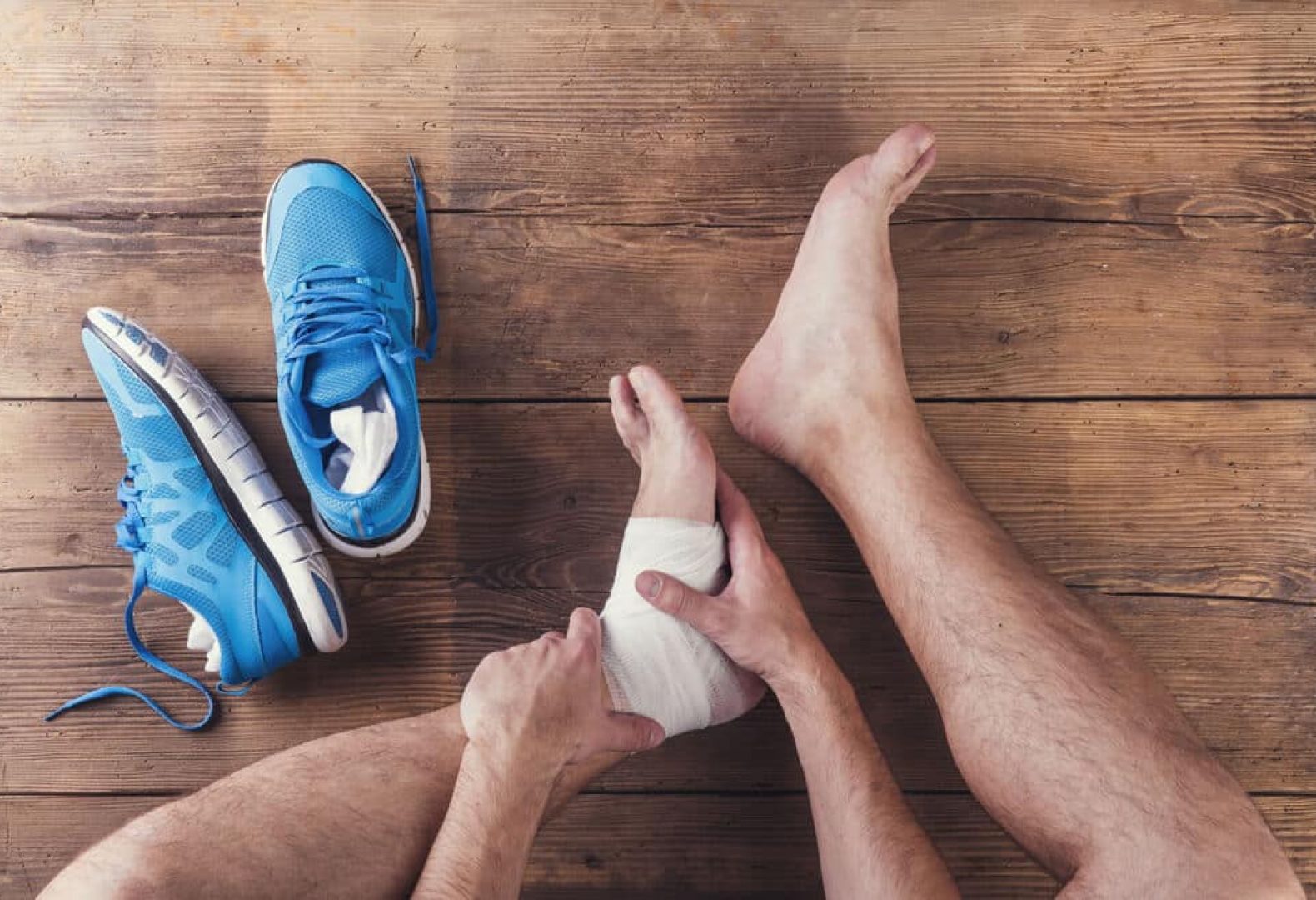 man with wrapped foot injuries sitting on wooden floor next to blue running shoes