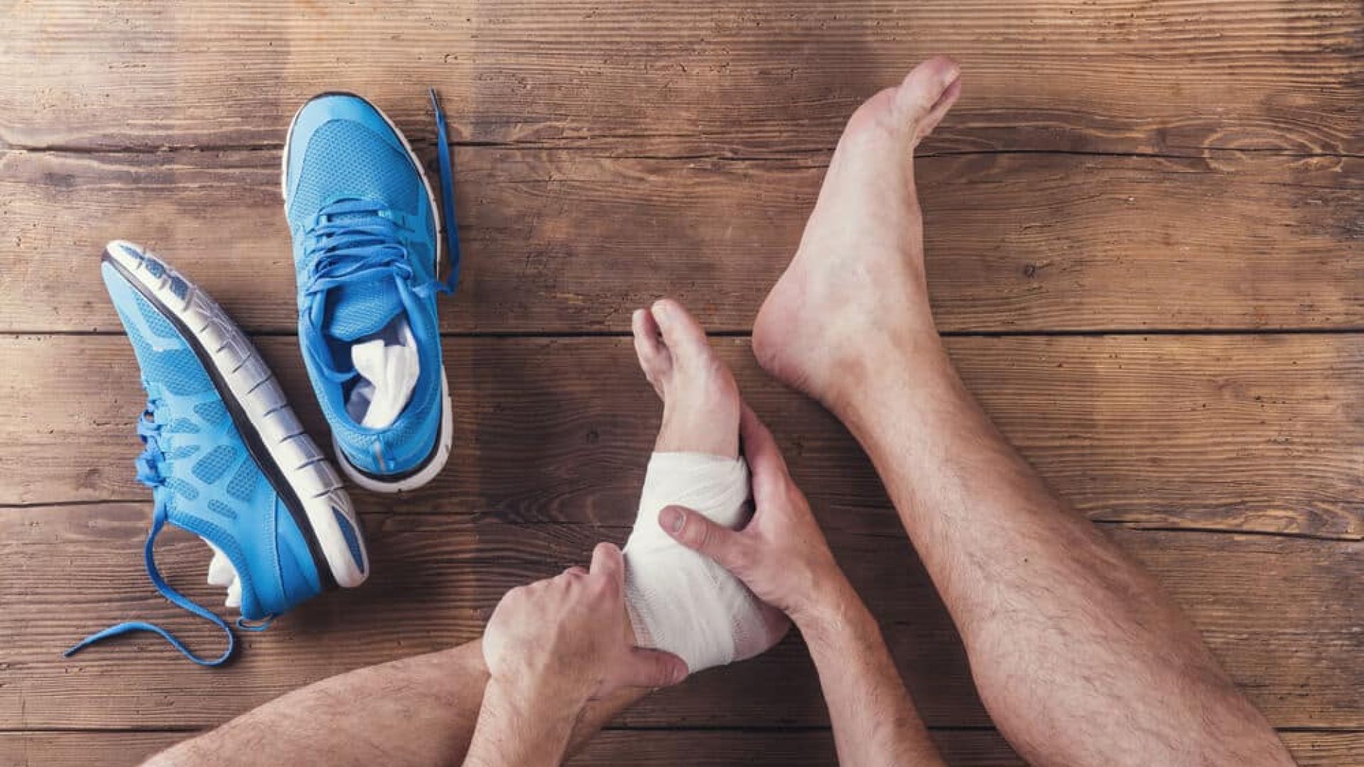 man with wrapped foot injuries sitting on wooden floor next to blue running shoes