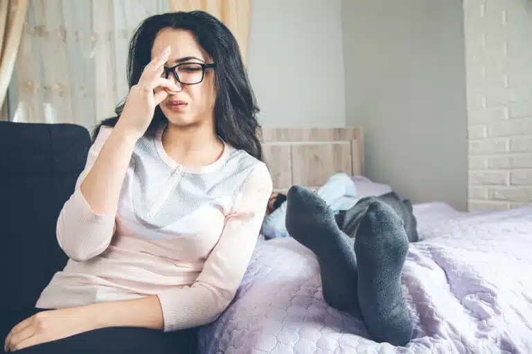 woman with glasses and dark hair pinching her nose due to stinky feet on bed next to her