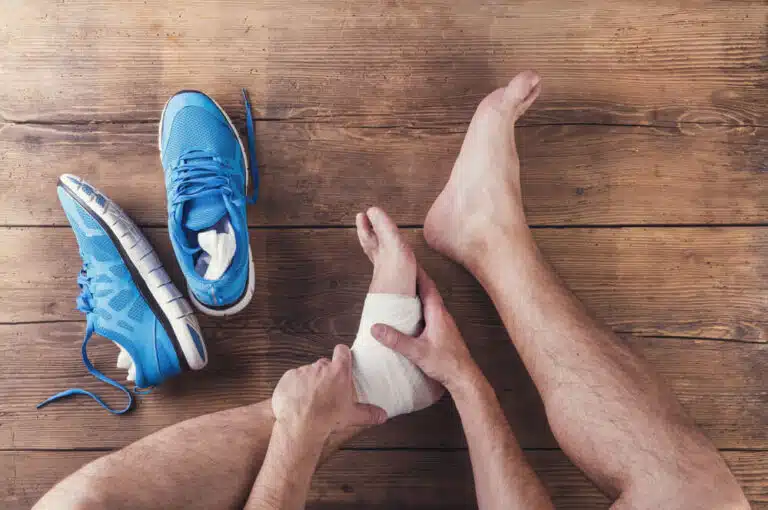 man with wrapped foot injuries sitting on wooden floor next to blue running shoes